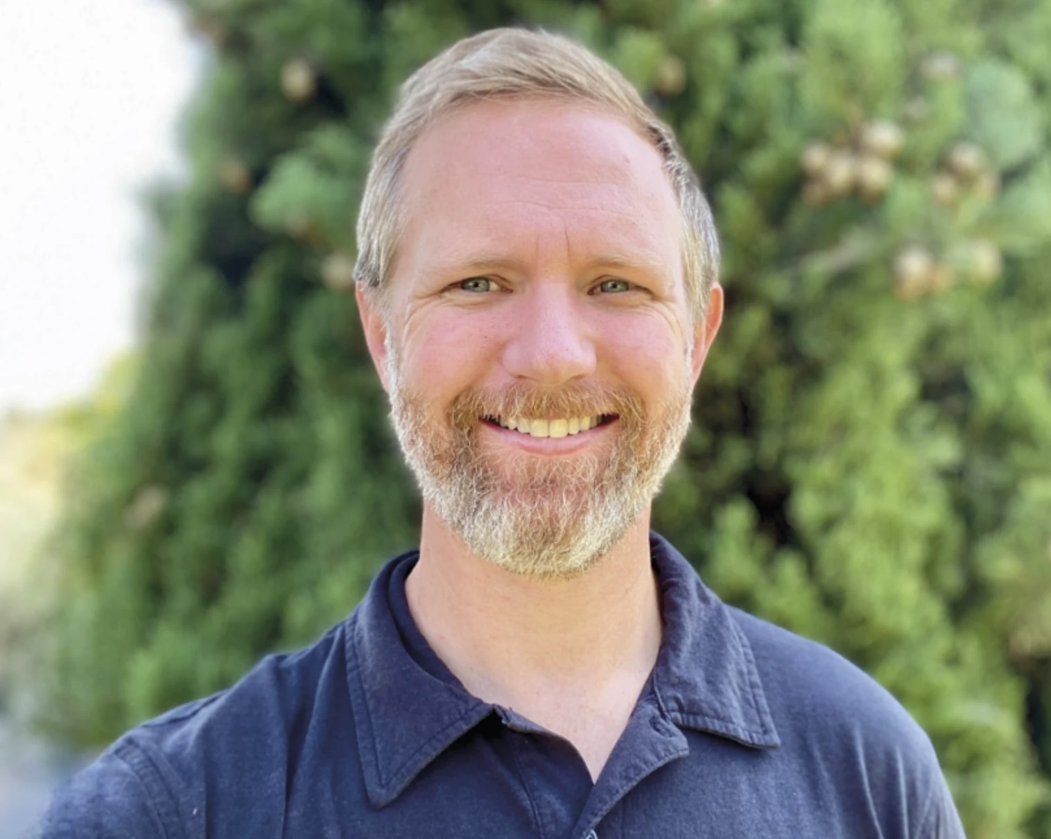 Portrait of a smiling bearded man in a navy polo shirt standing outdoors in front of greenery.
