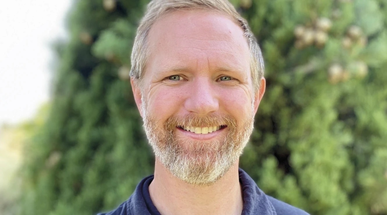 Portrait of a smiling bearded man in a navy polo shirt standing outdoors in front of greenery.