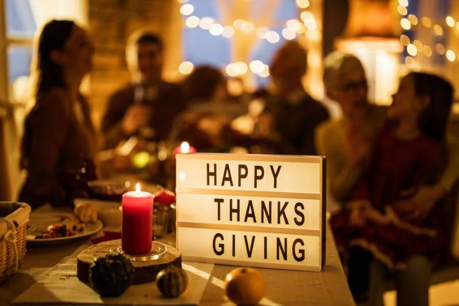 A lit red candle and string lights beside a light-box sign reading HAPPY THANKSGIVING on a table, with a group gathered for a holiday dinner in the background.
