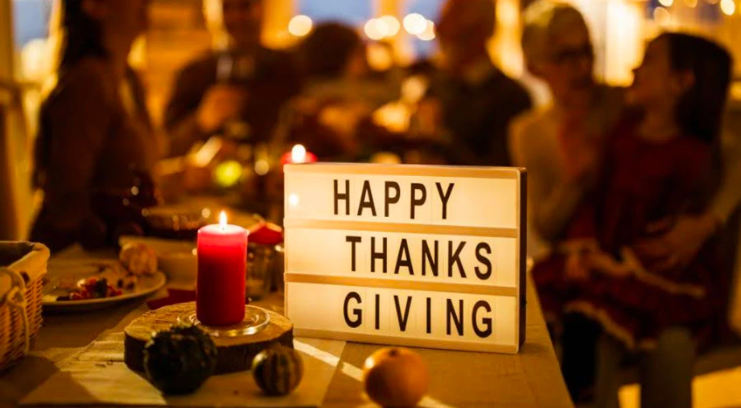 A lit red candle beside a light-box sign reading HAPPY THANKSGIVING on a table set for a holiday dinner, with blurred guests seated in the background.