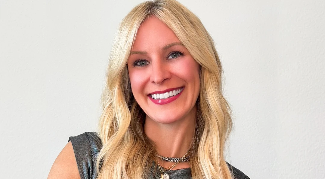 Portrait of a smiling blonde woman wearing a metallic top and layered necklaces against a plain light background.