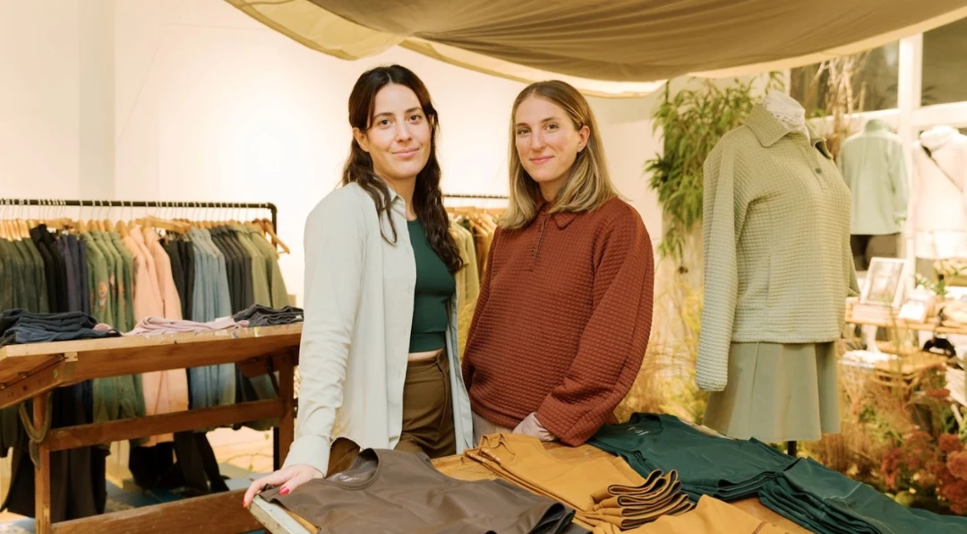 Two women standing together at a clothing display table filled with folded olive, green and mustard apparel under a canvas awning.