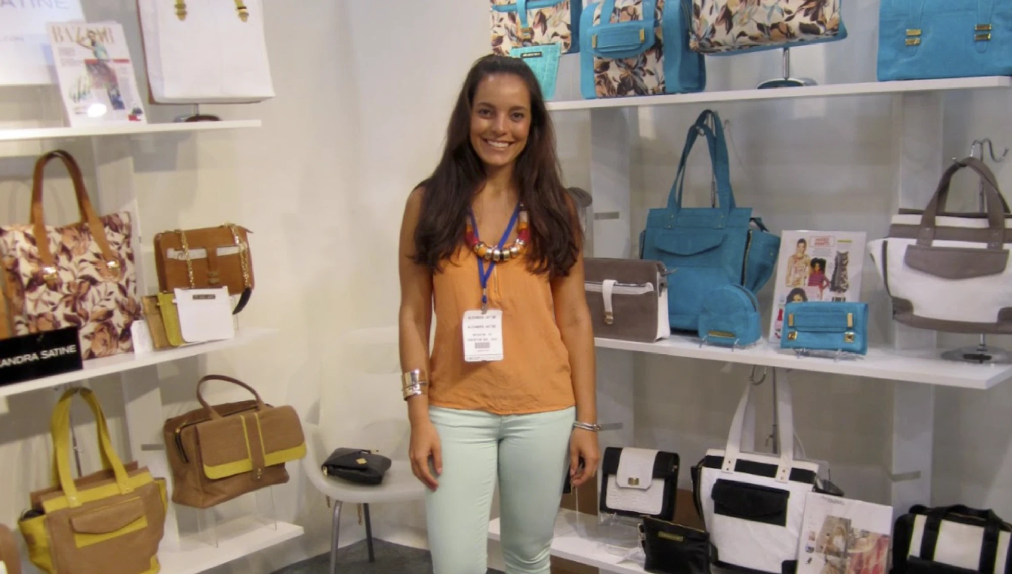 A smiling woman in a bright orange top standing in a booth surrounded by shelves of handbags on display.