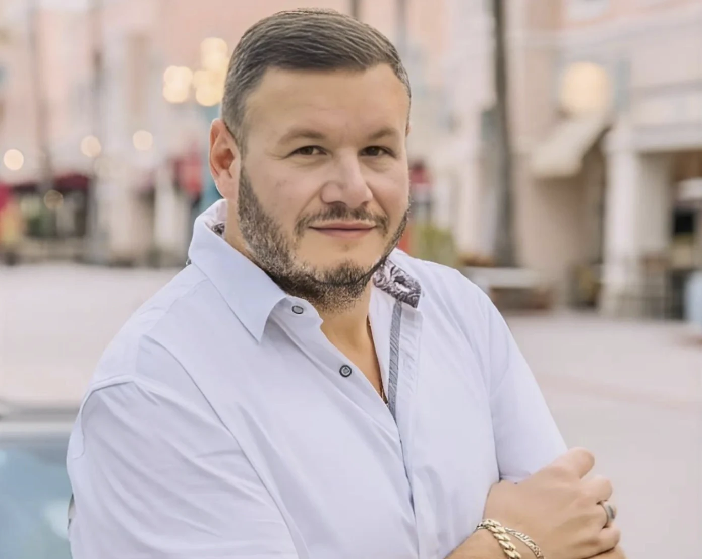 Portrait of a smiling man with a short beard wearing a light blue button-down shirt, standing on a city street at dusk.