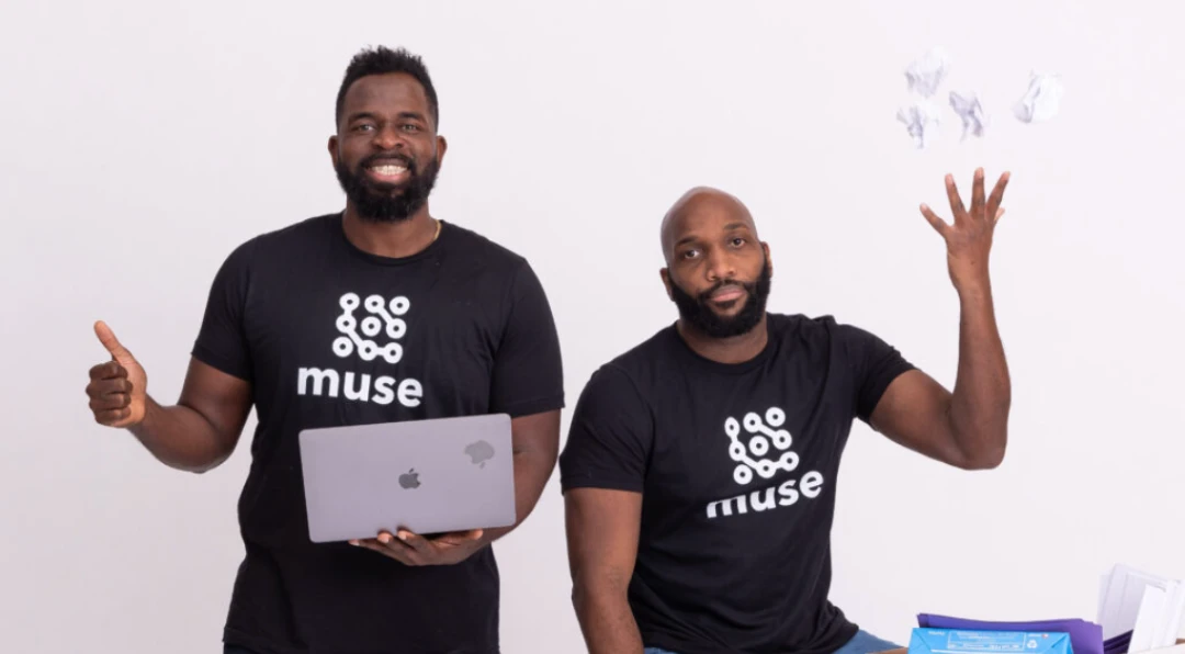 Two men in black Muse t-shirts posing together, one holding a laptop and giving a thumbs up, the other tossing crumpled paper in the air.