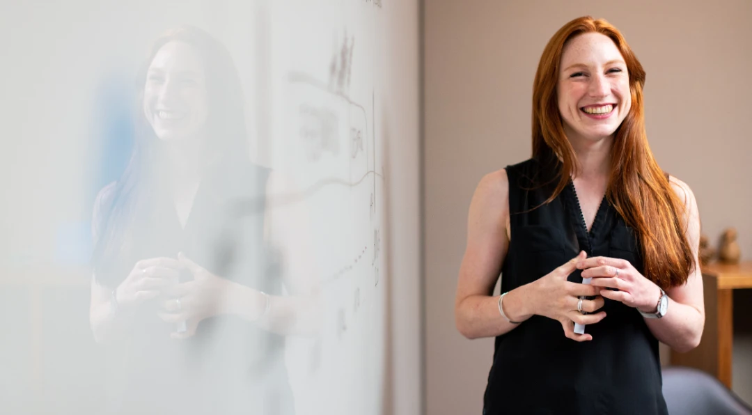 A red-haired woman in a black top laughing while standing beside a whiteboard with charts, her reflection visible in the glass.