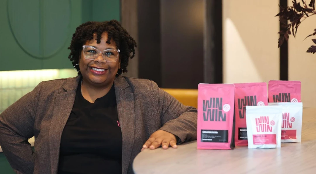 Nikisha Bailey smiling beside pink and white bags of Win Win Coffee on a café counter.