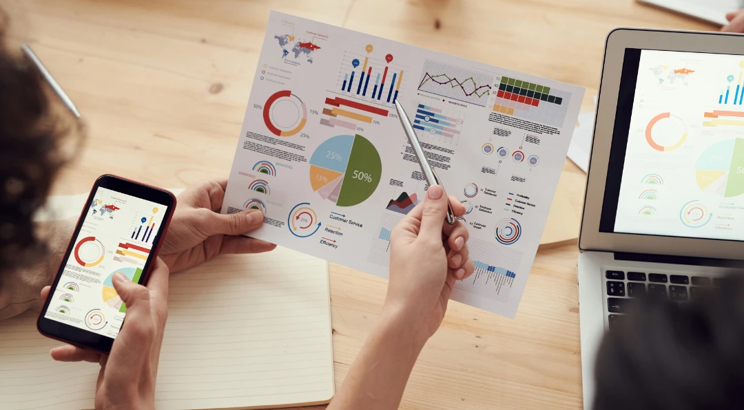 Overhead view of two people's hands holding a phone and a printed infographic with pie charts and bar graphs, beside an open laptop showing the same data.