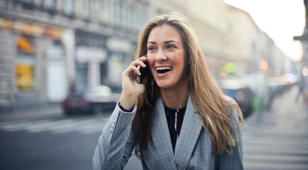 A smiling businesswoman in a plaid blazer talking on her phone while standing on a city street.