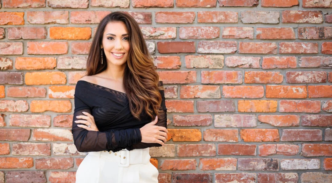 Portrait of a smiling woman with long dark hair, arms crossed, standing in front of a red brick wall.