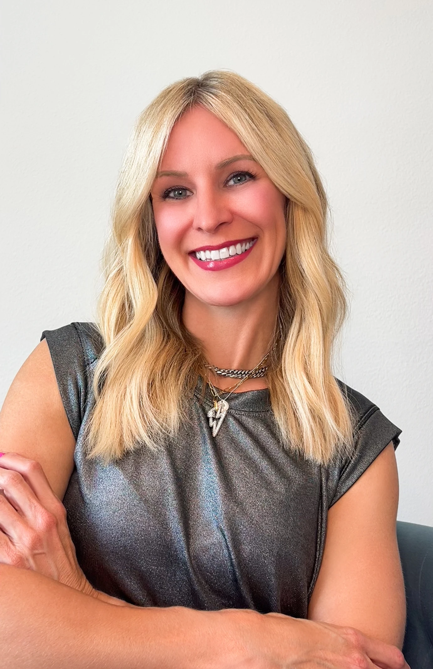 Portrait of a smiling blonde woman with her arms crossed, wearing a metallic top and layered necklaces against a plain light background.
