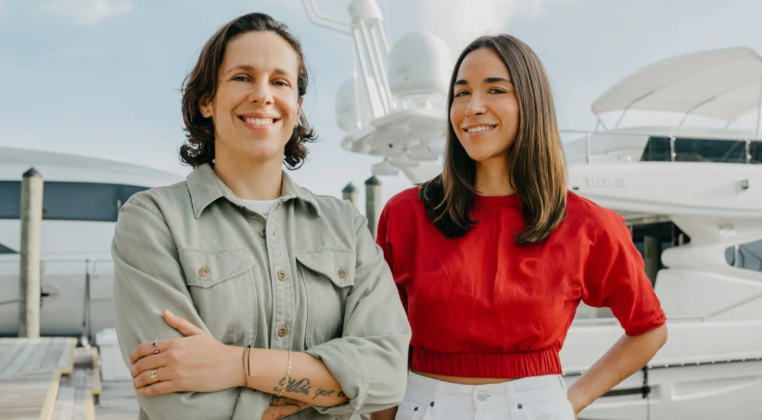 Two smiling women standing together on a dock in front of a large white yacht.