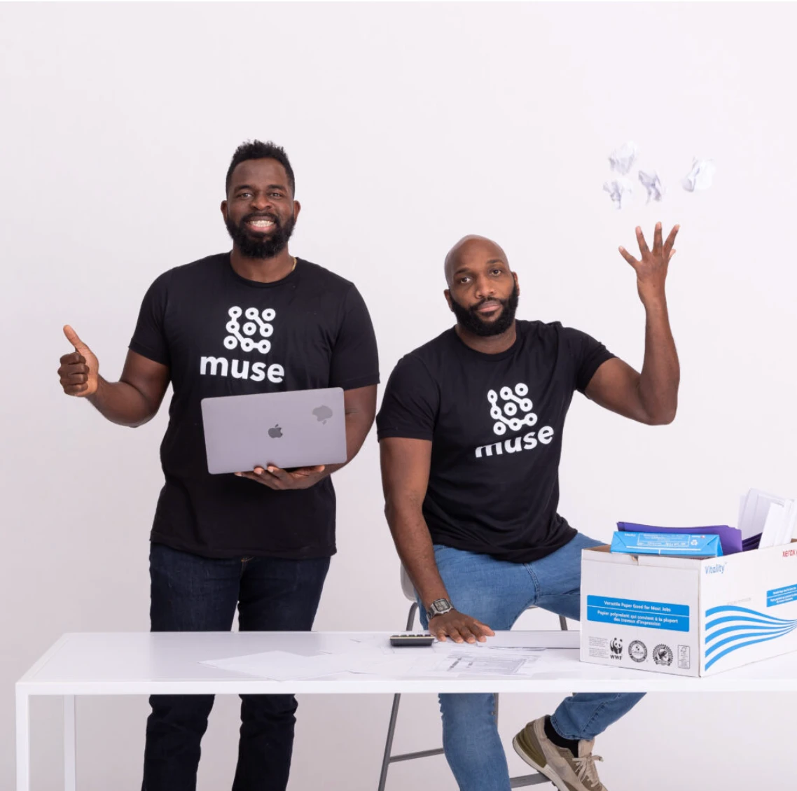 Two men in black Muse t-shirts posing together at a desk with a box of paper, one holding a laptop and giving a thumbs up, the other tossing crumpled paper in the air.