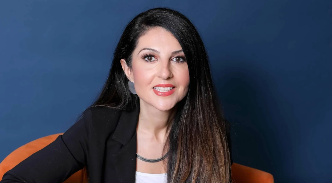 Portrait of a smiling dark-haired woman in a black blazer seated against a navy blue background.
